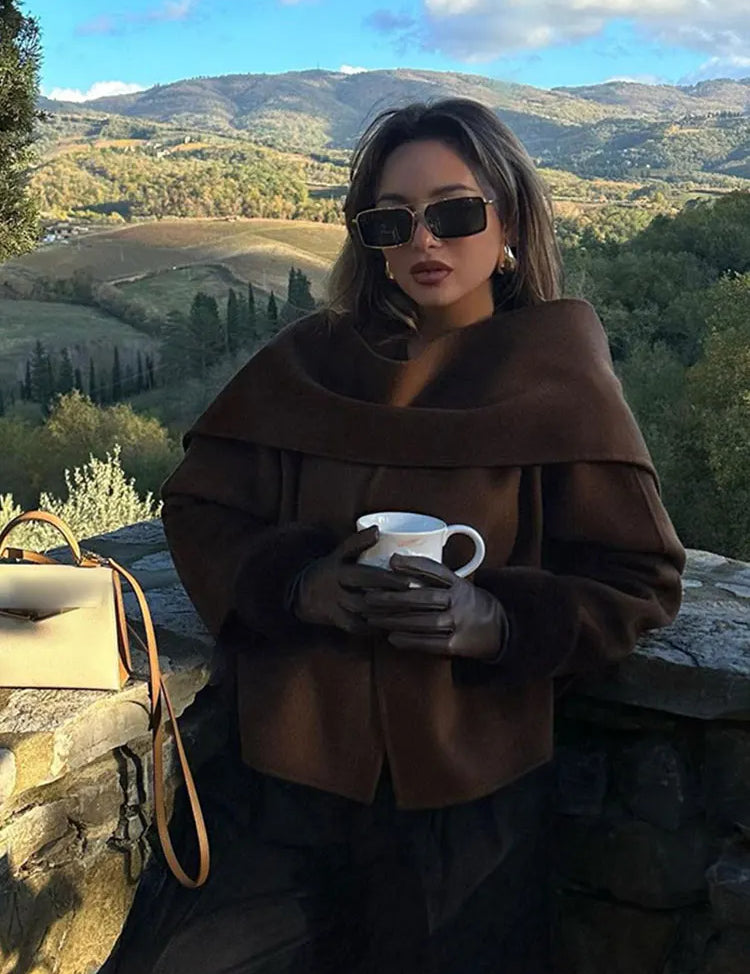 Donna con occhiali da sole e cappotto marrone, tiene una tazza di caffè su una terrazza panoramica in campagna. Paesaggio collinare autunnale sullo sfondo.