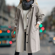 Cappotto beige donna, lungo e con bottoni, indossato su maglione bianco e sciarpa grigia, sfondo urbano sfocato, moda autunnale elegante.