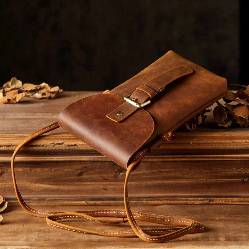 Borsa a tracolla in pelle marrone su tavolo di legno, design vintage, chiusura con bottone, ideale per accessori moda e stile retrò.