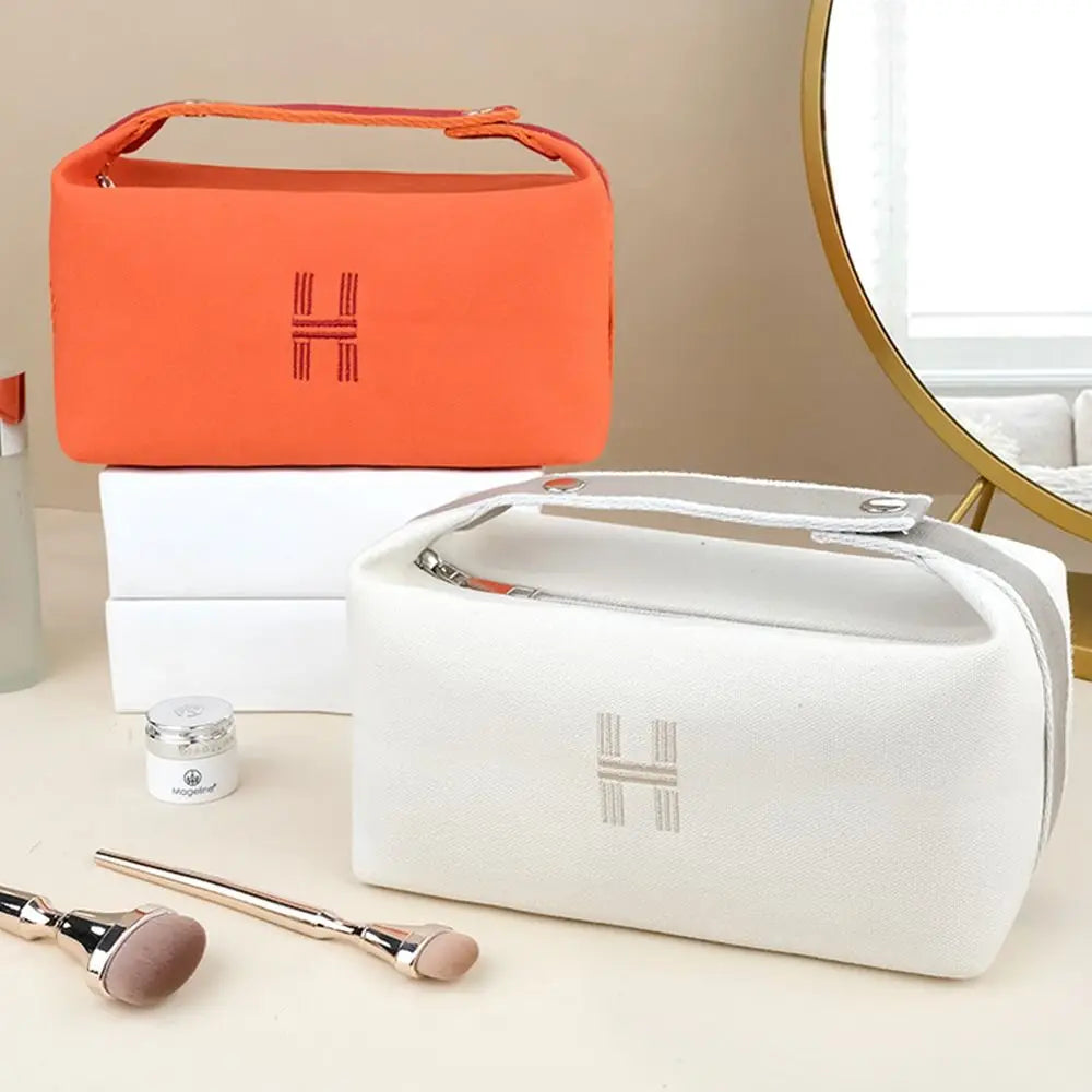 Borse cosmetiche in tessuto arancione e bianco con cerniera, logo ricamato, su tavolo con pennelli trucco. Elegante e funzionale per organizzare cosmetici.