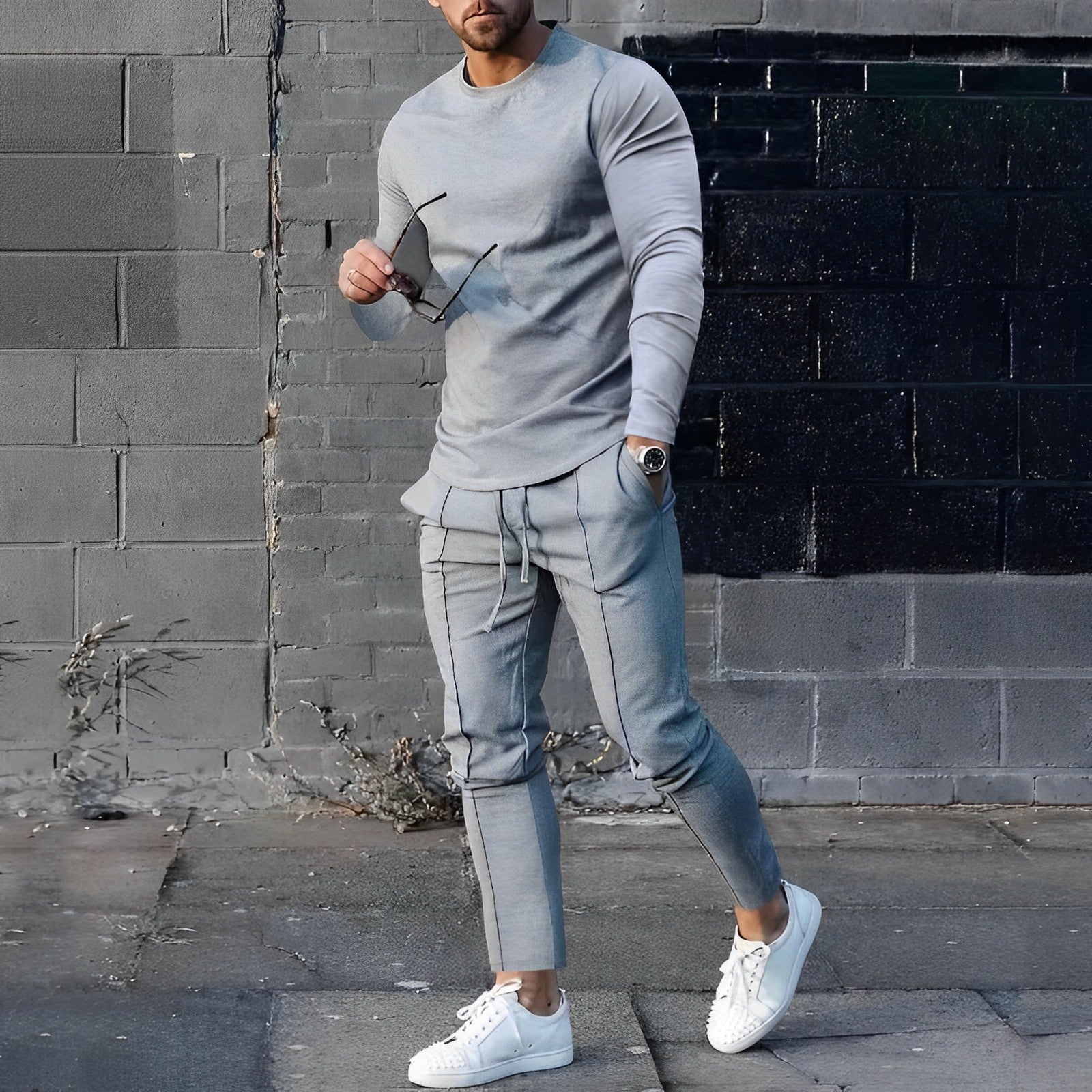 Uomo in abbigliamento casual grigio, indossa felpa e pantaloni abbinati, con occhiali da sole e scarpe bianche, su sfondo urbano.