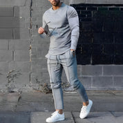 Uomo in abbigliamento casual grigio, indossa felpa e pantaloni abbinati, con occhiali da sole e scarpe bianche, su sfondo urbano.