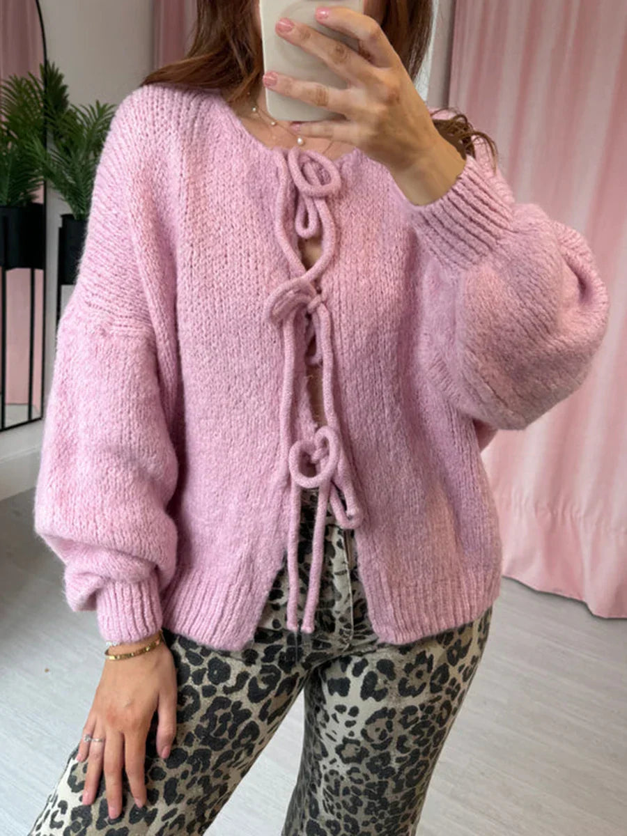 Donna con maglione rosa in lana con lacci, pantaloni leopardati, sfondo con tende rosa e pianta. Moda autunnale, abbigliamento casual chic.
