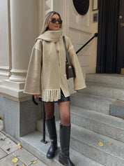 Donna con cappotto beige oversize e sciarpa abbinata, occhiali da sole, borsa a tracolla nera, stivali alti neri, su gradini di pietra. Moda autunnale elegante.
