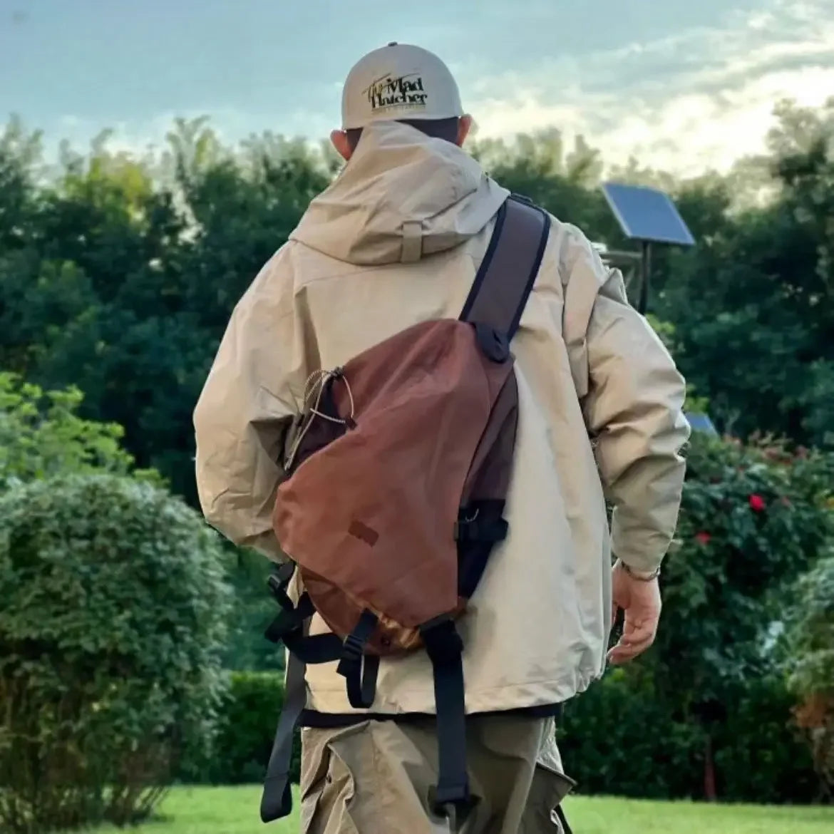 Uomo con giacca beige e zaino marrone in un giardino verde, indossa un cappello da baseball. Sfondo di alberi e cielo nuvoloso.