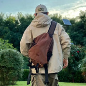 Uomo con giacca beige e zaino marrone in un giardino verde, indossa un cappello da baseball. Sfondo di alberi e cielo nuvoloso.