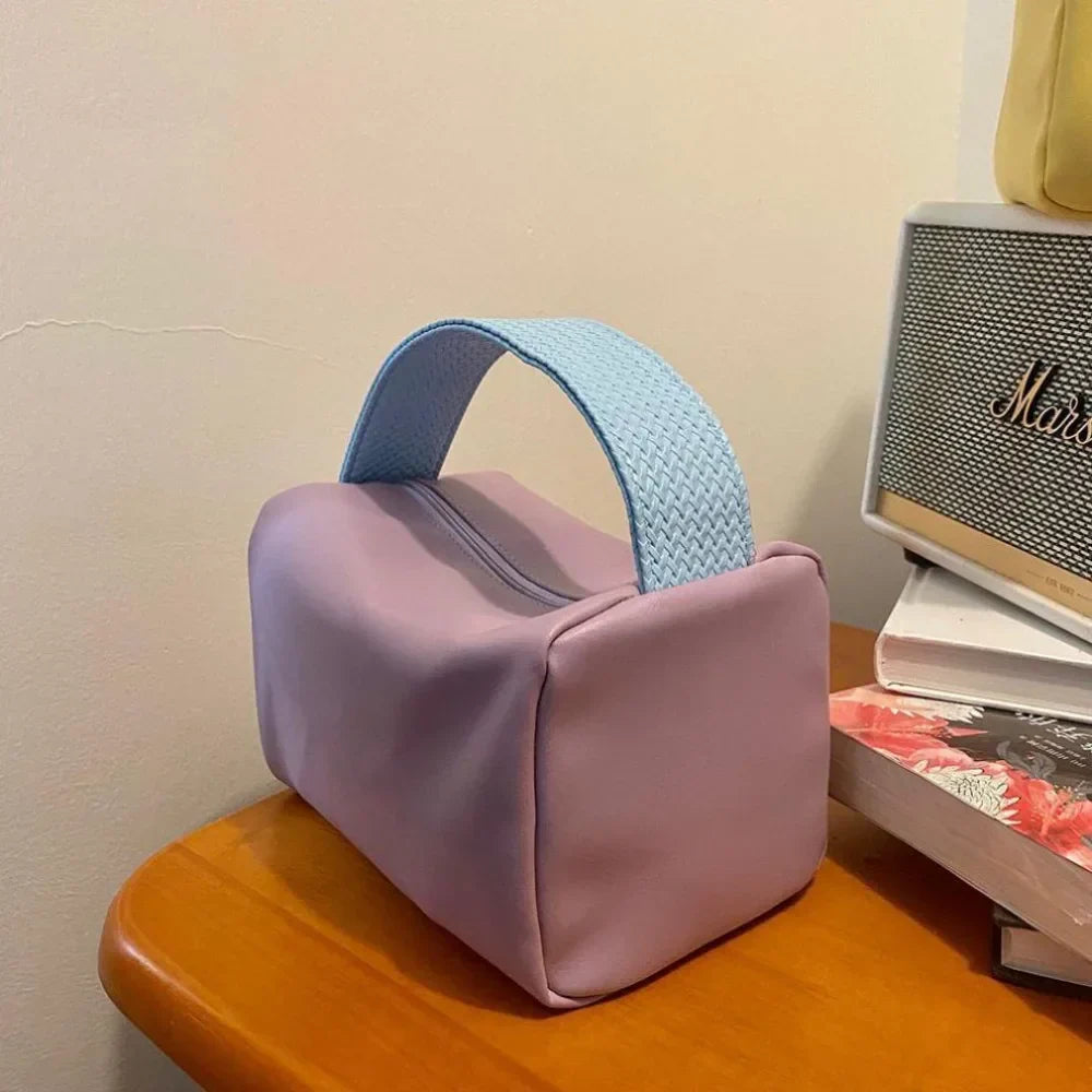 Borsa cosmetica viola con manico blu, design moderno, su tavolo in legno accanto a libri e radio vintage. Accessorio moda elegante e funzionale.