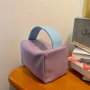 Borsa cosmetica viola con manico blu, design moderno, su tavolo in legno accanto a libri e radio vintage. Accessorio moda elegante e funzionale.