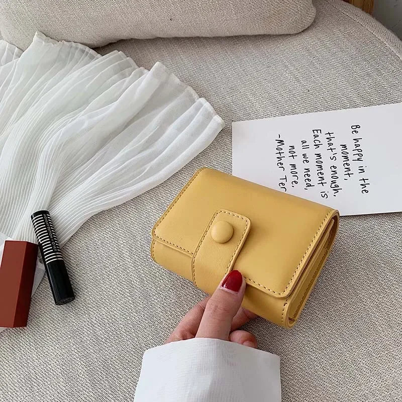 Portafoglio giallo in pelle su divano beige, accanto a rossetto nero e libro. Design compatto, chiusura a bottone. Accessorio moda elegante.