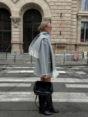 Donna con cappotto grigio e sciarpa abbinata, stivali neri alti, borsa nera, in piedi su strisce pedonali davanti a edificio storico. Moda autunnale urbana.