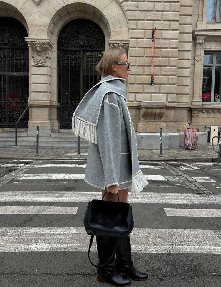 Donna con cappotto grigio e sciarpa abbinata, stivali neri alti, borsa nera, in piedi su strisce pedonali davanti a edificio storico. Moda autunnale urbana.