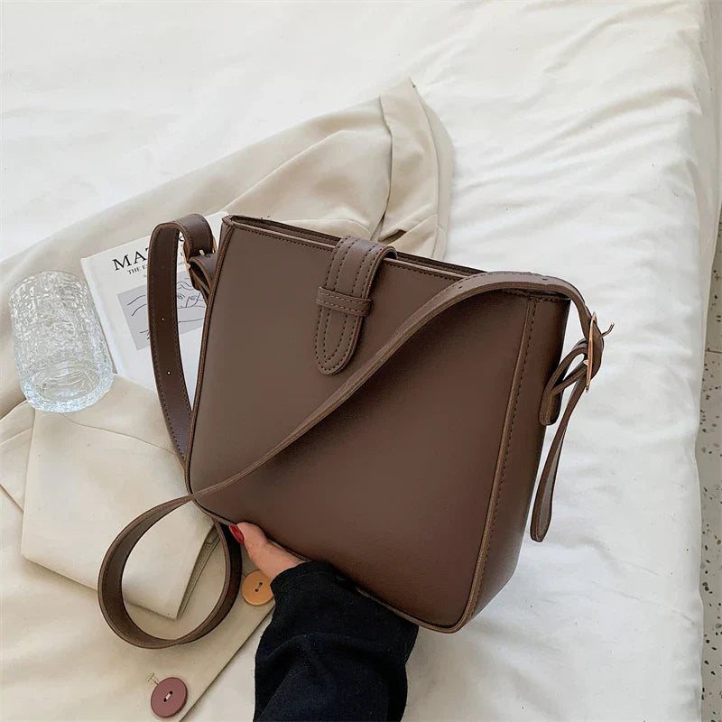 Borsa a tracolla marrone in pelle su letto bianco, con rivista e bicchiere d'acqua. Elegante accessorio moda donna, design minimalista.