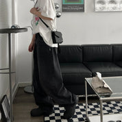 Utente in salotto moderno con divano nero, indossa jeans larghi neri, t-shirt bianca e borsa a tracolla nera. Arredamento minimalista.