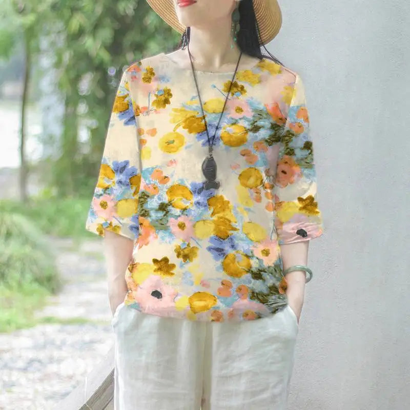Maglietta donna floreale, maniche corte, colori vivaci, stile casual estivo, indossata con pantaloni bianchi, accessoriata con collana e cappello.