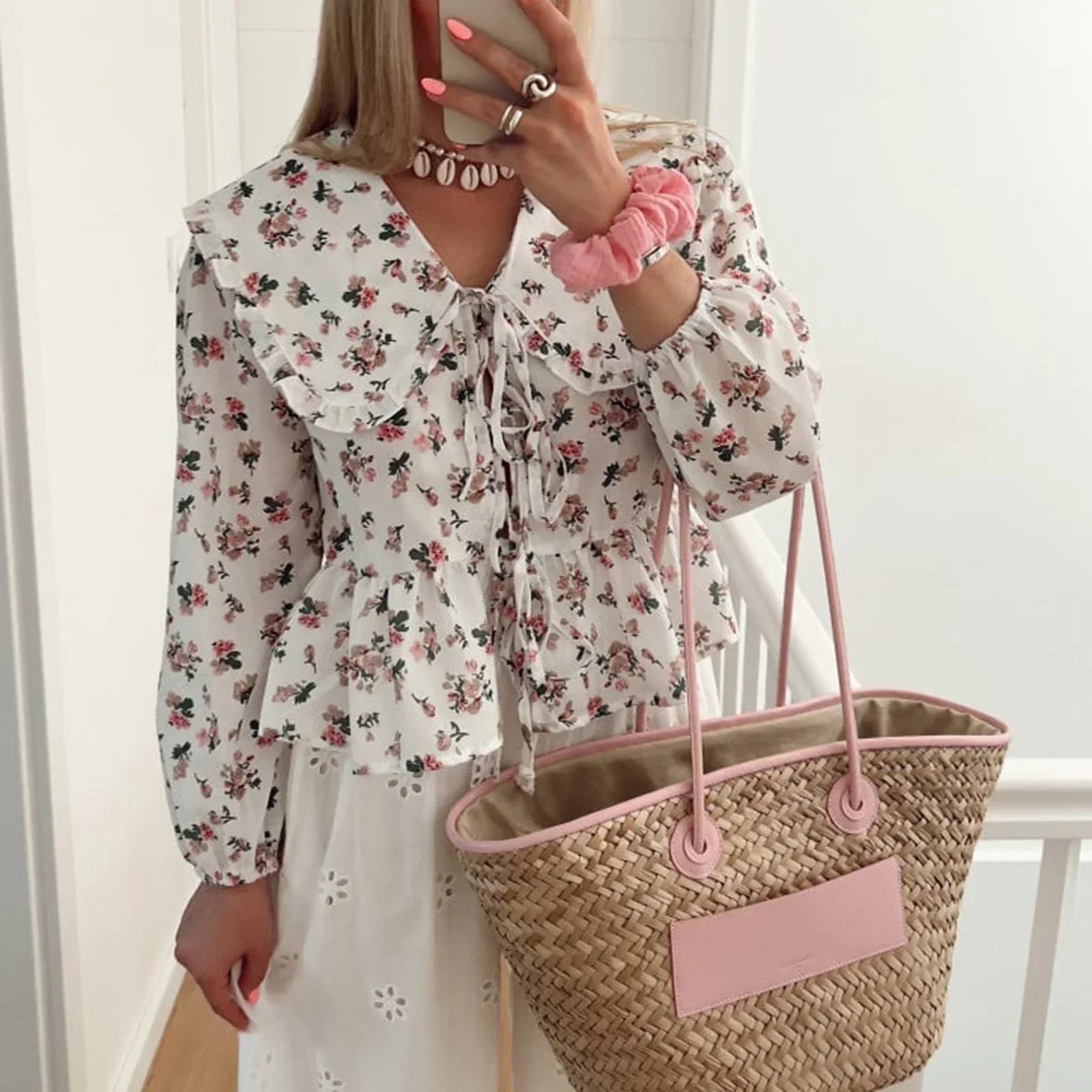 Donna con camicetta floreale bianca, gonna bianca traforata, borsa di paglia con manici rosa, bracciale rosa, collana di perle, unghie rosa.