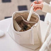 Borsa cosmetica beige aperta su tavolo bianco, contenente flaconi e scomparti organizzati. Mano femminile estrae spray. Accessori bellezza eleganti.