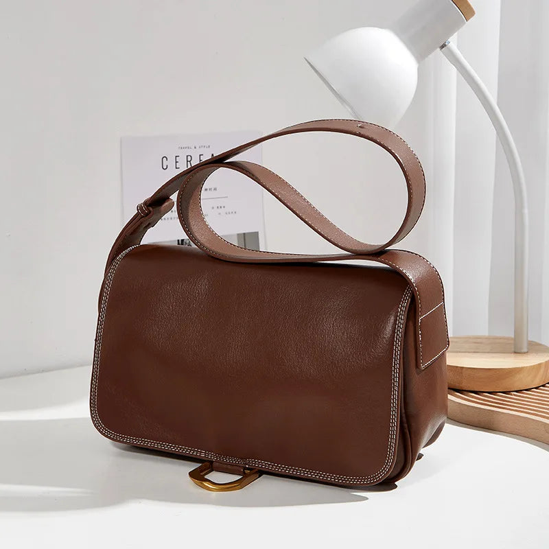 Borsa a tracolla in pelle marrone su tavolo bianco, con lampada moderna e libro. Elegante accessorio moda donna, design minimalista.