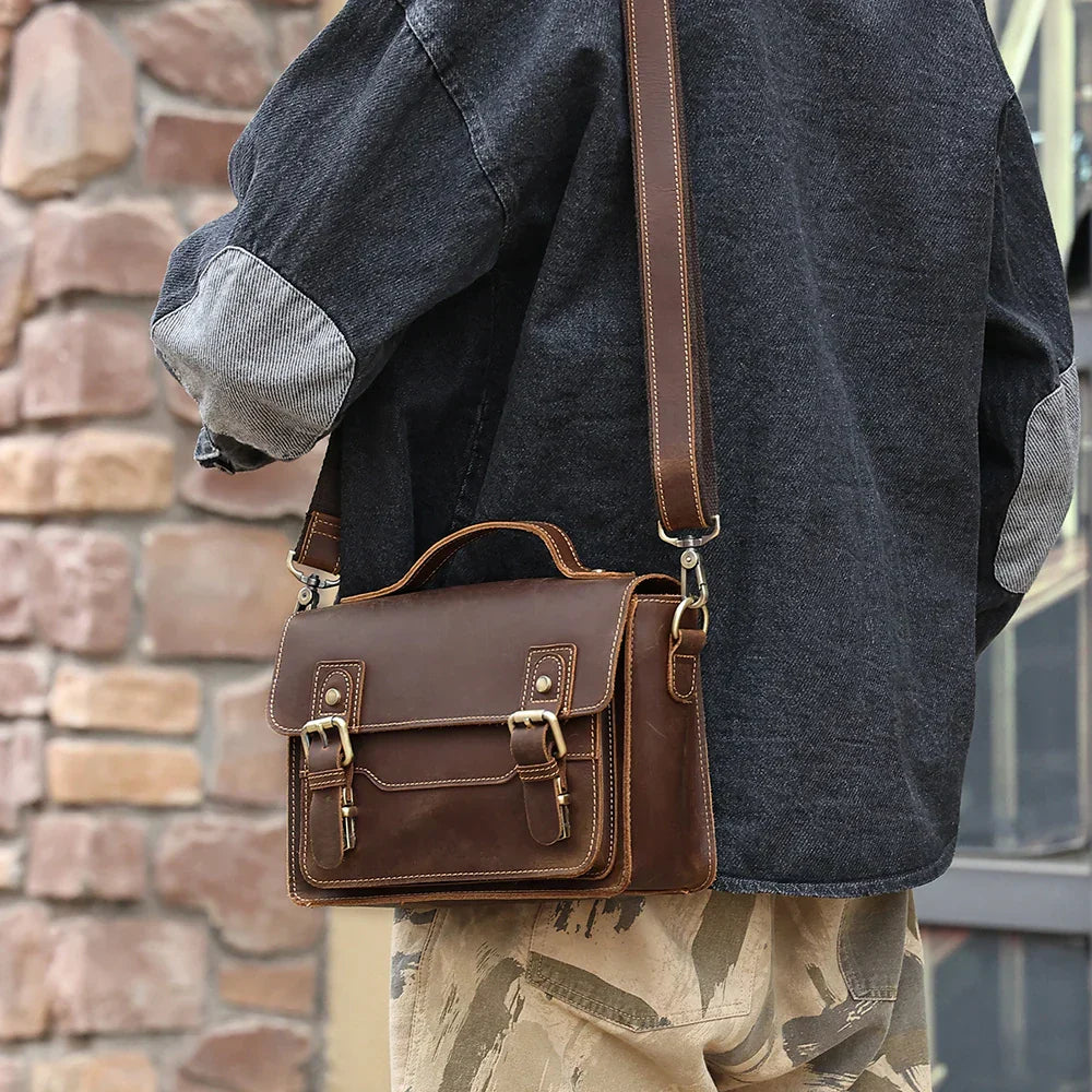 Borsa a tracolla in pelle marrone vintage, indossata da una persona con giacca grigia e pantaloni beige, davanti a un muro di mattoni.