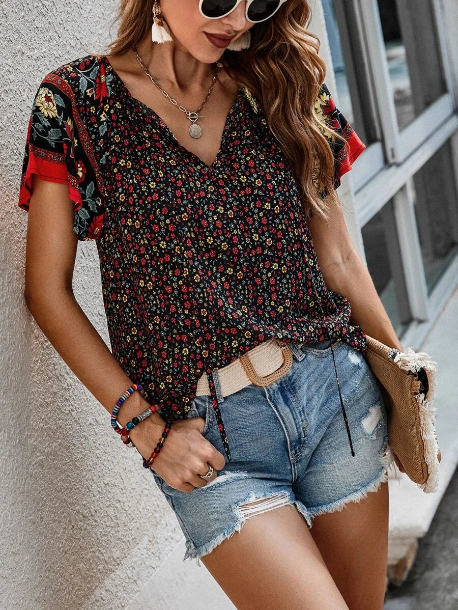 Donna con occhiali da sole indossa blusa floreale nera e rossa, shorts di jeans, accessori colorati, e borsa beige. Moda estiva casual femminile.