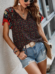 Donna con occhiali da sole indossa blusa floreale nera e rossa, shorts di jeans, accessori colorati, e borsa beige. Moda estiva casual femminile.