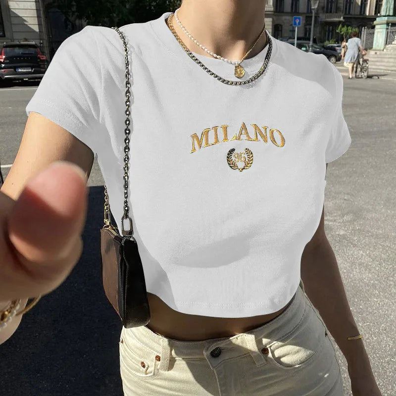 Donna con t-shirt bianca corta con scritta "Milano" in oro, collana dorata, borsa a tracolla nera, jeans chiari; moda estiva urbana.