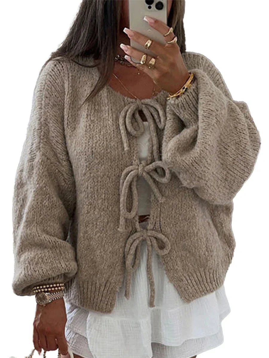 Donna con cardigan beige in maglia con fiocchi, maniche lunghe e ampie, sopra abito bianco. Stile casual chic, moda autunnale.