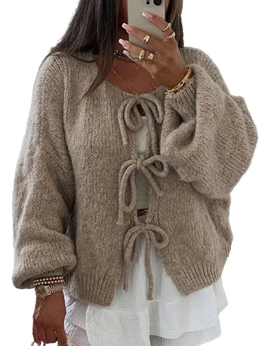 Donna con cardigan beige in maglia con fiocchi, maniche lunghe e ampie, sopra un abito bianco. Stile casual chic, moda autunnale.