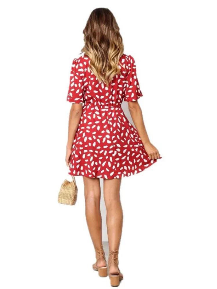 Abito estivo donna rosso a fantasia bianca, maniche corte, lunghezza sopra il ginocchio. Modella con borsa paglia e sandali. Moda casual elegante.