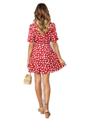 Abito estivo donna rosso a fantasia bianca, maniche corte, lunghezza sopra il ginocchio. Modella con borsa paglia e sandali. Moda casual elegante.