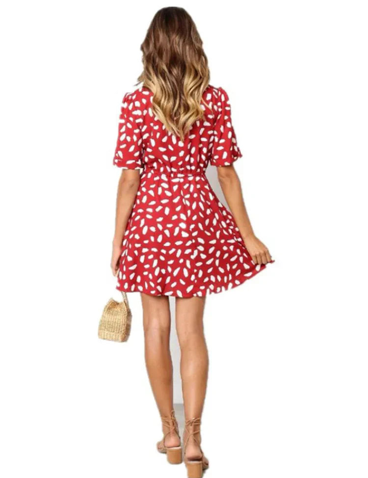 Abito estivo donna rosso a fantasia bianca, maniche corte, lunghezza sopra il ginocchio. Modella con borsa paglia e sandali. Moda casual elegante.