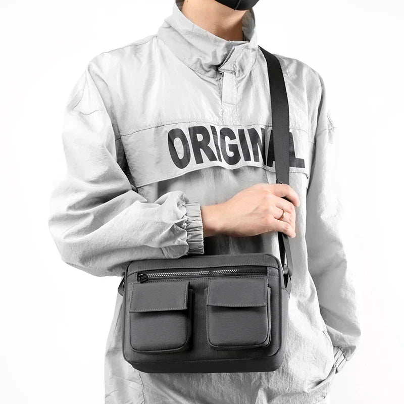 Borsa a tracolla nera unisex, design moderno con doppie tasche frontali, indossata su giacca grigia. Accessorio moda casual e funzionale.
