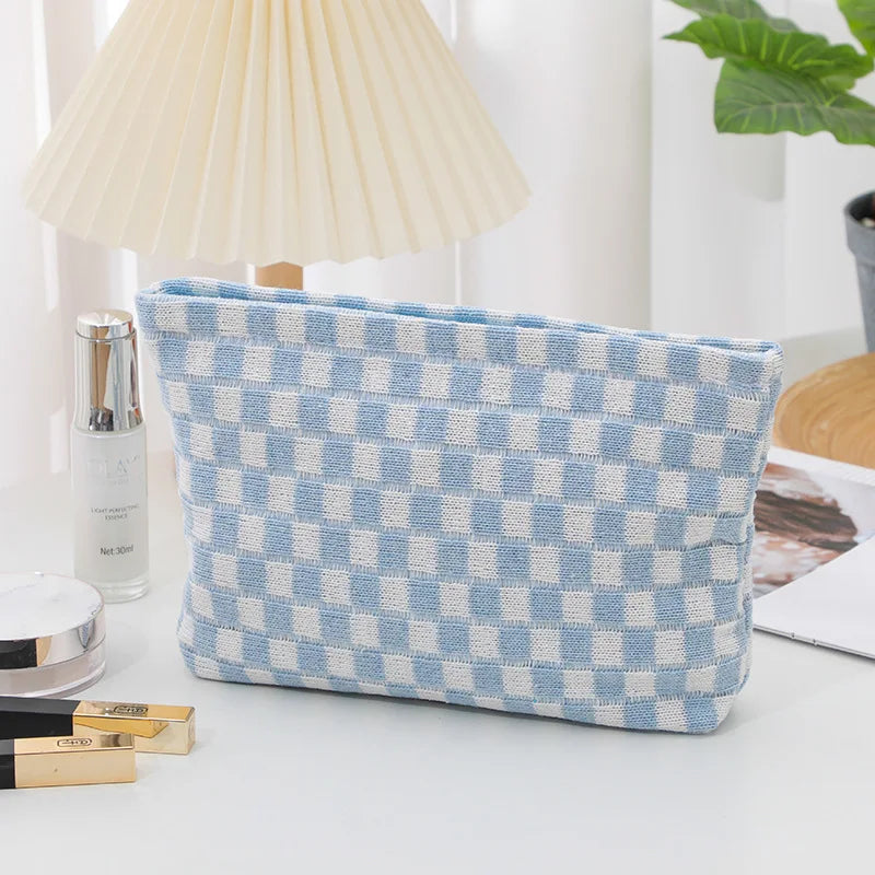 Pochette a scacchi blu e bianchi su tavolo bianco, accanto a lampada, piante e cosmetici. Accessorio moda elegante e pratico.