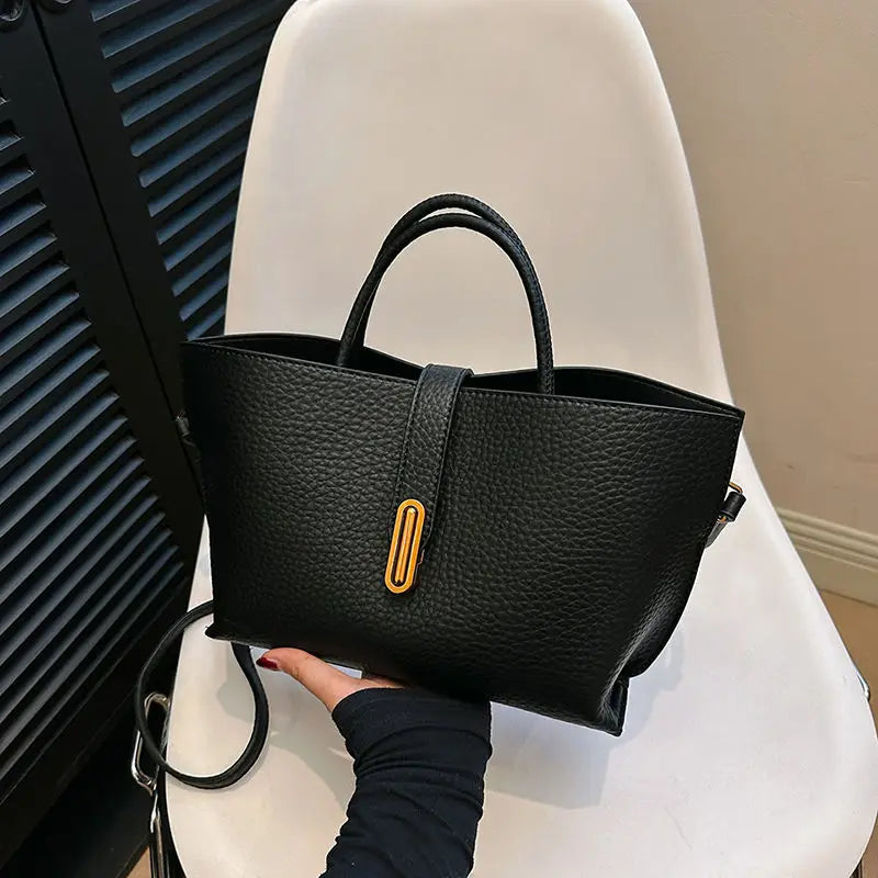 Borsa a mano in pelle nera con chiusura dorata, design elegante e moderno, su sedia bianca. Accessorio moda donna, stile sofisticato.