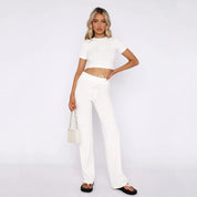 Donna in abbigliamento bianco: top corto e pantaloni a vita alta, borsa a catena, sandali neri. Moda estiva, look casual elegante.