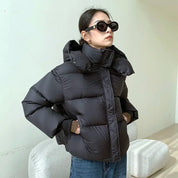 Giacca invernale donna nera, imbottita, con cappuccio, indossata da modella con occhiali da sole. Stile casual, moda autunno-inverno.