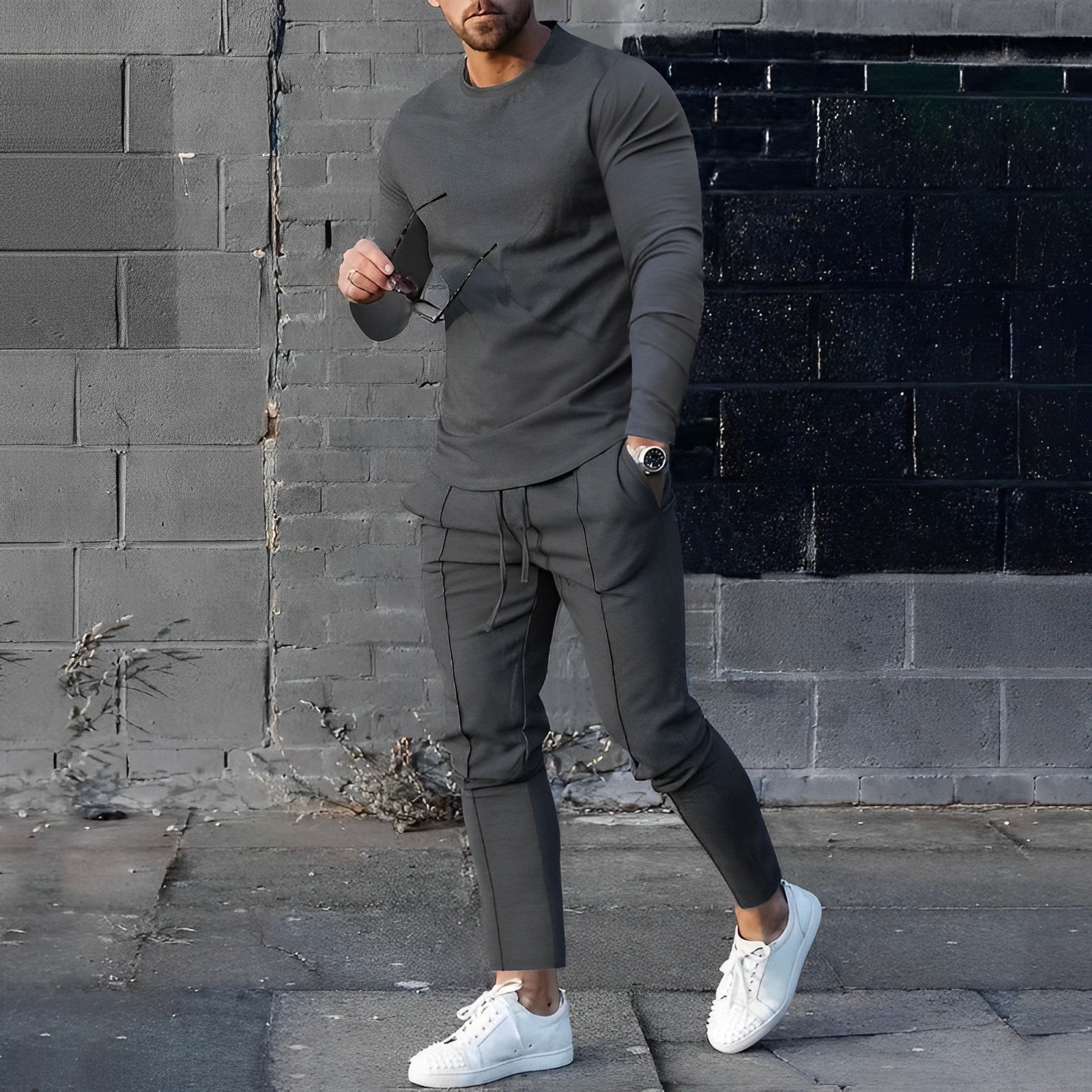 Uomo in abbigliamento casual grigio, con occhiali da sole e orologio, indossa sneakers bianche su sfondo urbano. Moda maschile moderna.