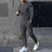 Uomo in abbigliamento casual grigio, con occhiali da sole e orologio, indossa sneakers bianche su sfondo urbano. Moda maschile moderna.