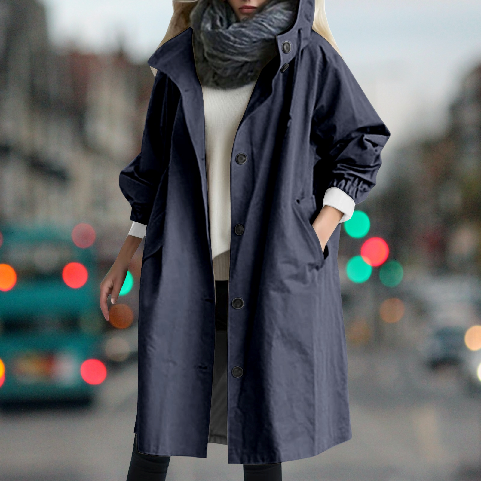 Cappotto blu scuro da donna, lungo e impermeabile, con bottoni e tasche, indossato su maglione bianco. Sfondo urbano sfocato.
