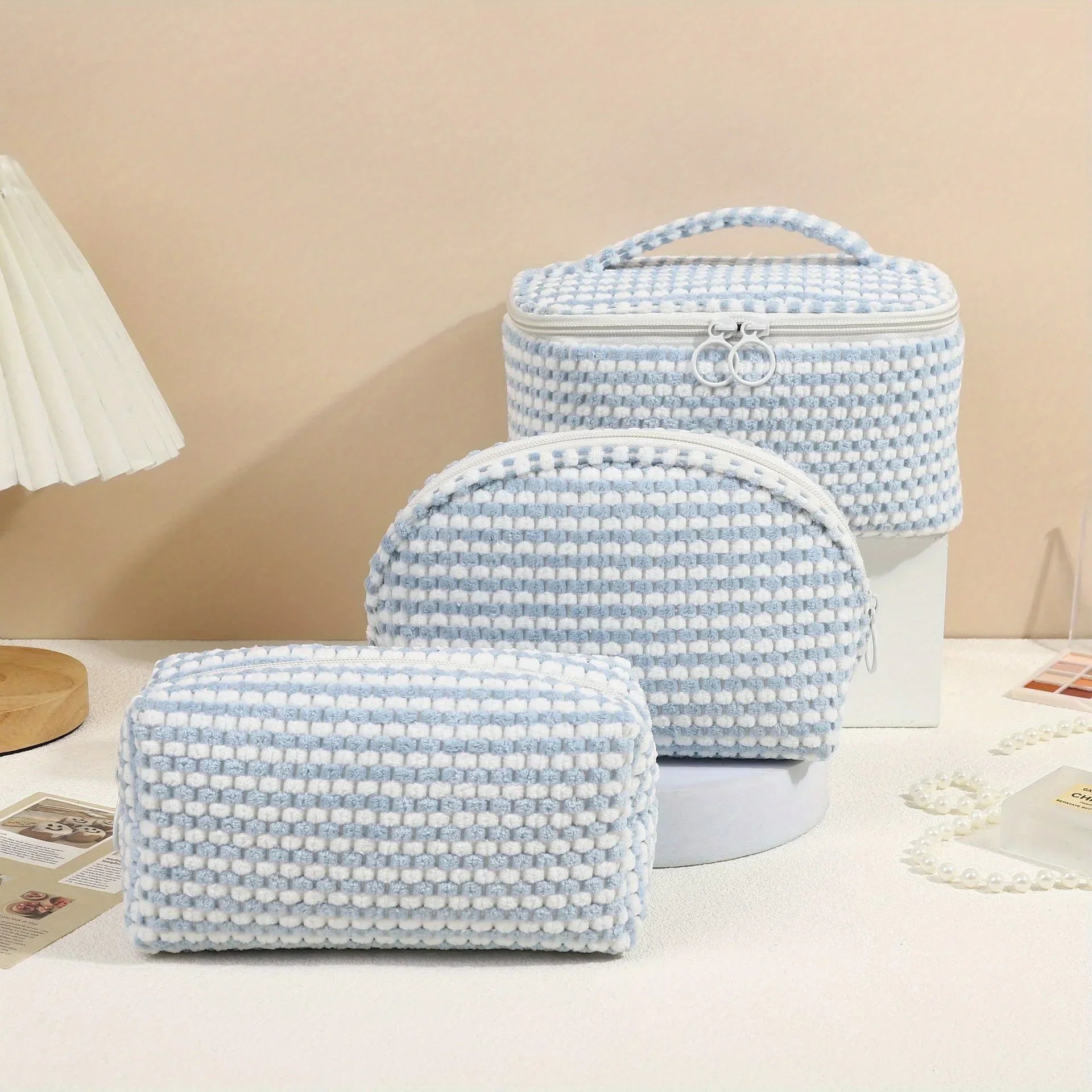 Set di tre borse da toilette in tessuto intrecciato bianco e azzurro, con cerniera e manico, su sfondo beige. Accessori da viaggio eleganti e pratici.