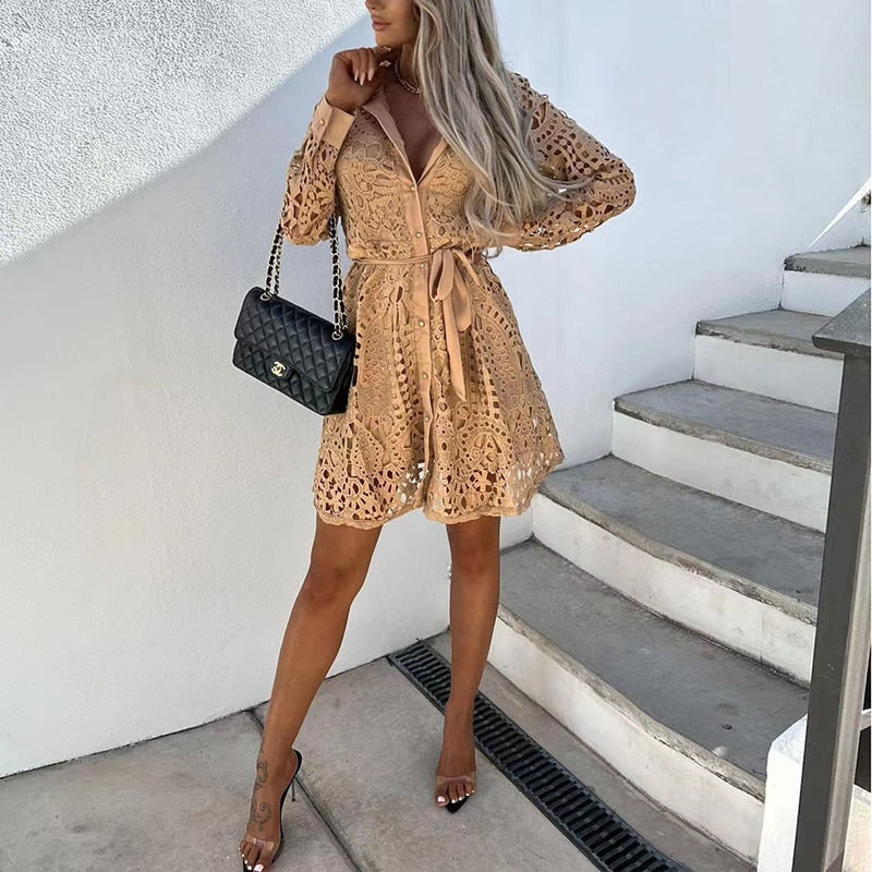 Donna con abito in pizzo beige, borsa a tracolla nera, sandali con tacco, posa su scala bianca. Moda estiva elegante, look chic e sofisticato.