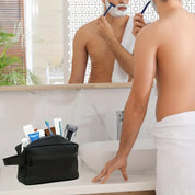 Uomo che si rade davanti allo specchio in bagno, accanto a un beauty case nero con articoli da toeletta come spazzolino, rasoio e dentifricio.