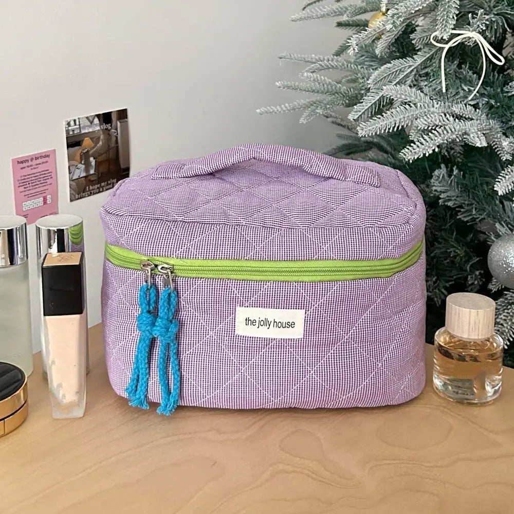 Astuccio cosmetico viola con cerniera verde, decorato con nappine blu, su tavolo vicino ad albero di Natale. Accessori trucco visibili.