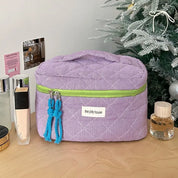 Astuccio cosmetico viola con cerniera verde, decorato con nappine blu, su tavolo vicino ad albero di Natale. Accessori trucco visibili.