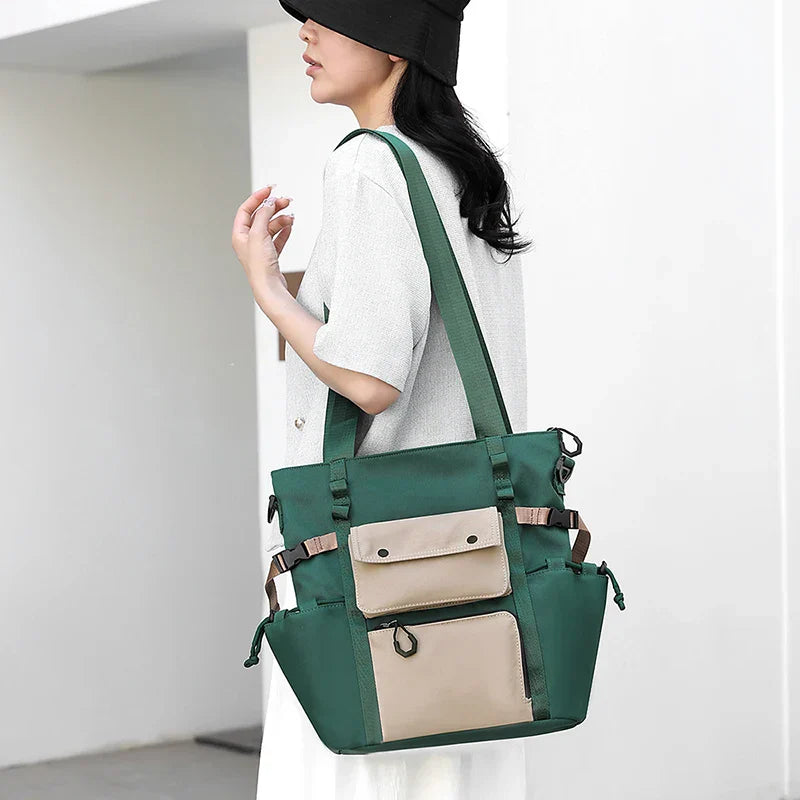 Borsa a tracolla verde con tasche beige, indossata da una donna in abito bianco e cappello nero. Design moderno, ideale per shopping e viaggi.
