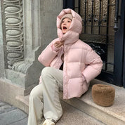 Giacca invernale rosa imbottita, donna seduta su gradino in città. Moda casual, cappuccio ampio, pantaloni chiari, stile urbano.