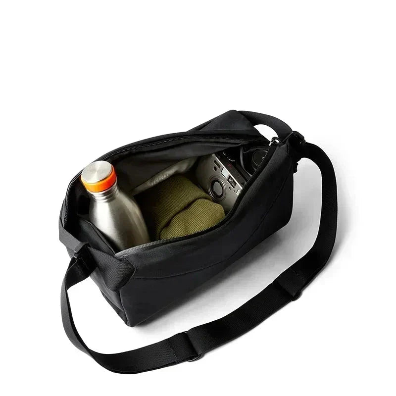 Borsa a tracolla nera aperta con bottiglia d'acqua, maglione verde e fotocamera all'interno, su sfondo bianco. Accessori viaggio, moda uomo.