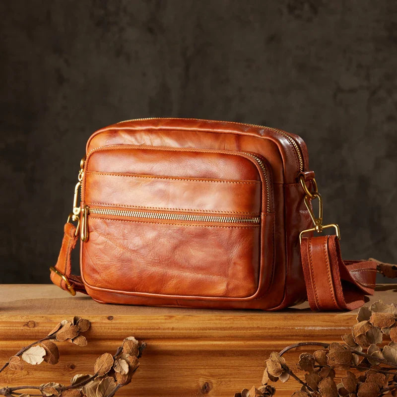 Borsa a tracolla in pelle marrone su tavolo di legno, design elegante con cerniere dorate, ideale per moda uomo e donna, accessorio di tendenza.
