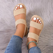 Sandali beige a doppia fascia su piedi femminili con smalto bianco, indossati con jeans e cavigliera turchese, su sfondo grigio peloso.