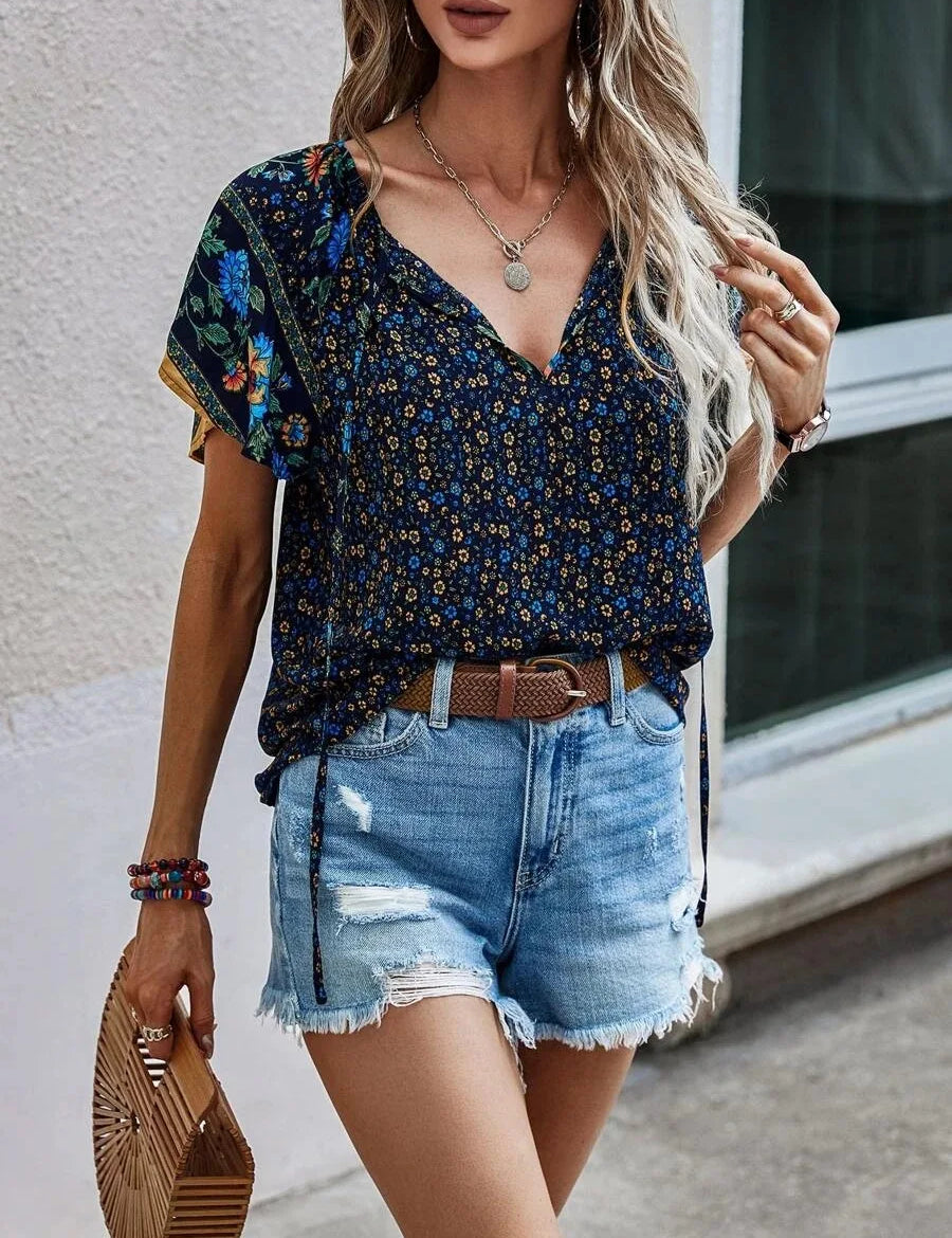 Donna con top floreale blu e shorts di jeans strappati, accessoriata con borsa in legno e bracciali colorati. Moda estiva casual e chic.