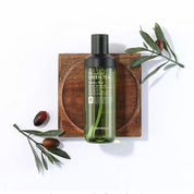 Bottiglia di tonico viso al tè verde TonyMoly su sfondo bianco, decorata con rami d'ulivo. Skincare coreana, idratante, naturale.